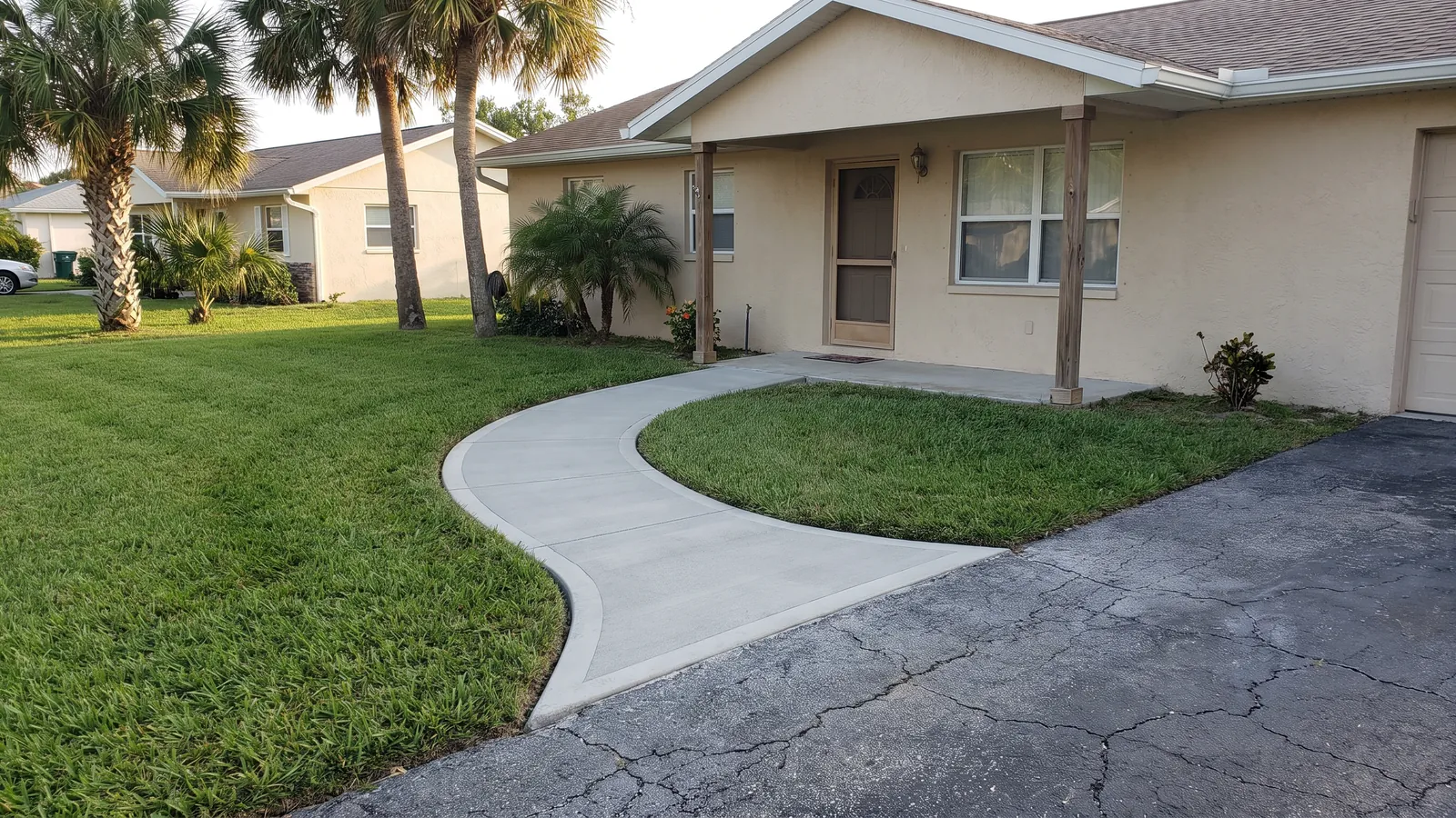 Curved broom-finished concrete front walkway with a smooth trowel border leading to the entry of a stucco block home with palms